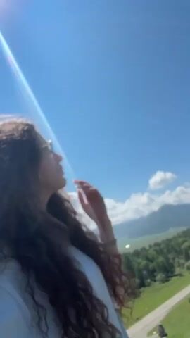 A woman with long, dark, curly hair and sunglasses is filming a video. She is standing outdoors on a grassy hill with a clear blue sky and mountains in the background. The camera pans to show a dirt path winding up the hill, with several people walking on it. The woman is smiling and appears to be enjoying the scenery.