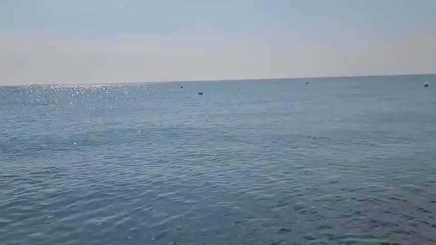The video shows a wide shot of the sea under a clear sky. The water is a calm blue, with gentle waves rippling across the surface. Sunlight glints off the water, creating sparkling highlights. In the distance, a few buoys are visible, as well as what appears to be a sailboat and a larger ship on the horizon. Towards the left side of the frame, a person is swimming in the water. The overall impression is of a peaceful day at sea.