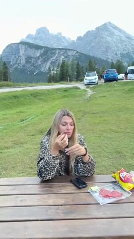 A woman with long blonde hair sits at a wooden picnic table outdoors, wearing a leopard-print jacket. She is holding a small object in her hands, seemingly examining it. In front of her on the table are a smartphone, a plastic container with some sliced food, and a bag of chips. The background features a grassy slope with trees and a large mountain range under an overcast sky. Several cars and a camper van are parked on a road in the mid-ground. The woman appears to be eating or preparing to eat.