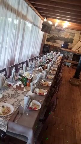 The video shows the interior of a restaurant or dining area set for a large gathering. The camera pans across the room to display the layout and decor. There is a long table set for what appears to be a formal meal, with plates, cutlery, glasses, napkins, and bottles of beverages. There are multiple wooden chairs arranged along both sides of the table. In the background is a large fireplace with logs. 

The room has a rustic ambiance with wooden beams on the ceiling and stone or brick accents on the walls. The word "Nazdrave" is painted on the wall.

The additional seating area has wooden booths and tables set with similar arrangements as the long table. The room looks ready for guests.