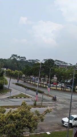 This is a top-down, medium-angle shot of a road intersection with trees and buildings in the background. The sky is overcast. On the left, a white car is driving away from the camera. In the middle of the frame, a motorcyclist is turning right. On the right, a white SUV is parked. There are two streetlights in the middle of the intersection with Malaysian flags attached to them.