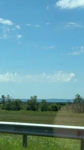The video captures a daytime view from a moving vehicle, likely a car, traveling along a road in a rural area. A grey metal guardrail runs along the bottom of the frame, separating the road from a wide, green grassy field. Beyond the field, power lines and poles are visible. In the distance, a hazy outline of mountains or hills stretches across the horizon. The sky is bright blue with scattered white clouds, indicating clear weather. The overall scene depicts an open, expansive countryside landscape, consistent with the provided location context of Kazazov, Russia. No people or visible text are present.
