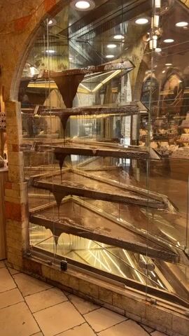 A mesmerizing tiered chocolate fountain display is captured within a glass enclosure, showcasing thick, melted chocolate cascading downwards. The chocolate flows from a funnel-like structure at the top, over several slanted, flat shelves, creating individual waterfalls that drip into the shelf below. The rich, dark brown chocolate pools on each level before continuing its descent.

The display is framed by a decorative, stone-like or brick-patterned archway, adding an elegant and somewhat rustic touch. Bright internal lighting illuminates the flowing chocolate, highlighting its texture and movement. Reflections on the glass enclosure reveal the interior of a shop, likely a confectionery or chocolate store, with shelves displaying various products and additional lighting fixtures. An arched architectural detail is visible in the background reflection, consistent with the location of San Gimignano, Italy. Briefly, a person can be seen walking past in the reflection on the right side of the frame. Faint, illegible text is visible on some of the shop shelves in the background, and the word "CRUZ" or "CROS" can be partially seen on the chocolate structure at the very top. The sound of dripping chocolate is audible throughout the video.
