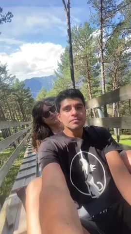 The video shows a couple riding what appears to be a gravity-powered sled or cart on a wooden track through a forest. The man in the foreground is filming a selfie-style video. He is wearing a black t-shirt with a white circular graphic. He smiles and makes a peace sign with his fingers. Behind him, a woman with long brown hair and sunglasses is seated. She leans her head on his shoulder and smiles. The track is elevated and made of wood, with a wooden railing on the left side. Tall pine trees surround the track, and a mountainous landscape is visible in the background under a cloudy blue sky. The lighting suggests it is daytime.