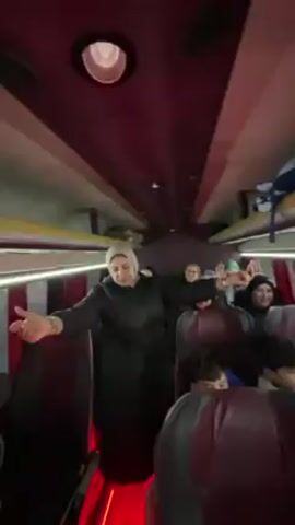 Inside a moving bus, a woman wearing a hijab and a black coat dances in the aisle. She faces toward the back of the bus, moving her arms and clapping her hands. Several people, including women and boys, sit in the seats, some smiling and raising their hands. The bus has burgundy seats and dark red lighting near the floor. A few bags are stowed on overhead racks.