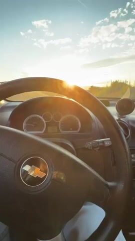 This is a first-person perspective video taken from inside a car. The camera is pointed forward through the windshield. The sun is setting in the distance, casting a warm glow and lens flare across the scene. The sky is blue with scattered white clouds. The dashboard of the car is visible in the foreground, including the steering wheel with a Chevrolet logo, instrument cluster, and part of the car's interior. A road stretches out ahead, leading towards a small structure in the distance. The surrounding environment appears to be rural or semi-rural, with vegetation and some fencing visible. The car is in motion.