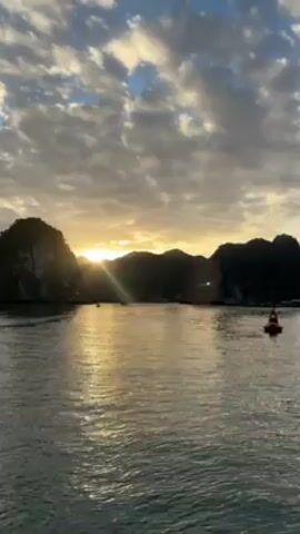 The video displays a scenic view of a body of water with islands in the background under a cloudy sky. The sun is setting, creating a golden glow that reflects off the water. A red navigational buoy is visible in the water. The landscape is characterized by dark, silhouetted islands. Two people speaking are faintly audible, but their identities and specific words are indiscernible. The camera appears stable, suggesting it is handheld or placed on a steady surface.