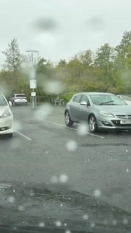 The video shows a rainy parking lot. A silver Vauxhall Astra is parked in a space, and other cars are parked nearby. Raindrops are visible on the windshield of the vehicle from which the video is being recorded. Trees and a gray sky can be seen in the background.