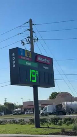 The camera is angled up at a RaceTrac gas station sign that displays the price of diesel and regular gas. Diesel is listed at $5.19 per gallon and regular is $3.76 per gallon. The sign is black with green numbers for diesel and red numbers for regular. There are power lines visible above the sign and buildings and tanker trailers in the background. The sky is blue.