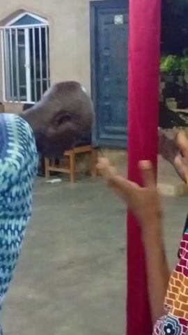 The video depicts three individuals inside a building. The individual in the foreground, a woman, is clapping her hands and singing. She is wearing a colorful dress with a dark head wrap. To her left stands a man in a blue patterned suit, facing away from the camera. Behind the woman is an older individual, dressed in black, also facing toward the singing. A red pole stands prominently in the center of the frame. The building has a concrete floor and a simple roof structure. In the background, through a doorway, there is a glimpse of foliage and a building. Blue plastic chairs are scattered around.