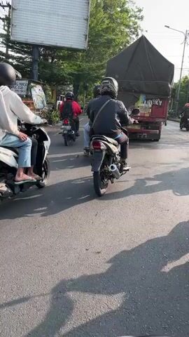 The video shows a street scene with several motorcycles and a truck. The motorcycles are moving in the same direction, with riders wearing helmets. The environment appears to be a city street with buildings and trees in the background. The lighting suggests it is daytime, possibly late afternoon due to the long shadows. One motorcycle in the foreground has two passengers, one driving and one riding on the back. The truck is in the distance, partially obscured by a tree.