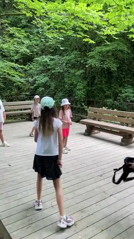 The video captures a group of children playing on a wooden deck surrounded by lush green foliage. Two girls in the foreground, one in a white t-shirt and black skirt, the other in a pink t-shirt and pink shorts, are engaged in some sort of game. They are both wearing hats, one light blue and the other white. Another girl, in a white t-shirt and black skirt, stands further back on the right. A boy, wearing patterned shorts, is positioned in the center of the frame, facing the girls. He appears to be holding a black backpack.

The deck is furnished with wooden benches, suggesting a park or recreational area. The background is dominated by dense green trees and vegetation, indicating an outdoor setting likely in a natural environment. The sunlight filtering through the leaves suggests it is daytime, and the weather appears pleasant.

The children's body language suggests they are actively participating in a game, with movements indicating throwing, catching, or some form of playful interaction. There are no discernible emotions explicitly conveyed, but the context implies a lighthearted activity. No visible text is present in the video. The location context provided is Izmaylovka, Russia, which aligns with the natural, possibly wooded, environment depicted.