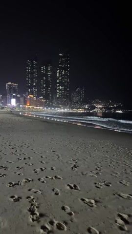 The video shows a sandy beach at night. In the background, a cityscape with tall buildings is visible, their lights illuminating the dark sky. The buildings appear to be quite densely packed. The beach is covered in footprints. The waves are gently lapping at the shore, with their foamy edges reflecting the city lights. The overall impression is one of urban development meeting the natural environment.