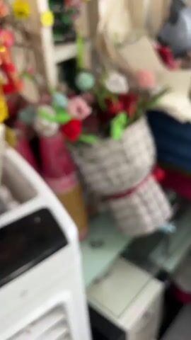 The video shows a cluttered indoor environment. In the foreground, a white air cooler with ventilation slots is visible on the left. A glass-topped table occupies the center, with a bouquet of artificial flowers in a textured gray wrap placed on it. The flowers are a mix of red, pink, white, and blue, with green stems and leaves. To the left of the bouquet are two tall, cylindrical objects wrapped in pink and gold paper, possibly candles or decorative items. In the background, a white shelf unit is visible, decorated with green tinsel and small red and green ornaments. Various other objects are piled around the shelf, including what appears to be a grey helmet and folded fabrics. On the table, a small ginger and white kitten is visible to the right of the bouquet, facing left. A second ginger and white kitten is partially visible behind it. The lighting suggests an indoor, possibly domestic setting.