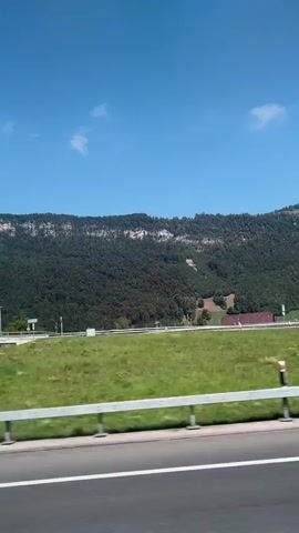 The video begins with a view of a bright blue sky with a few white clouds. A dense, green forest covers the side of a mountain range that rises in the background. In the foreground, there is a green field bordered by a white guardrail. A section of a concrete overpass or bridge comes into view, with its underside casting shadows on the ground below. The video then returns to the same scenic view of the mountain, forest, and field. The camera is moving, suggesting it is filmed from a vehicle.