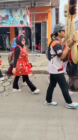 A group of people are walking down a street. The people are wearing various outfits. Some people are wearing red dresses with white trim, and black leggings, and headscarves. Others are wearing more traditional clothing. One person is wearing a black and blue patterned shirt and black pants. There is a shop in the background with a sign that says "SEGARWANITA" and other text in Indonesian. There is also a flag visible. The ground is paved. It appears to be daytime.