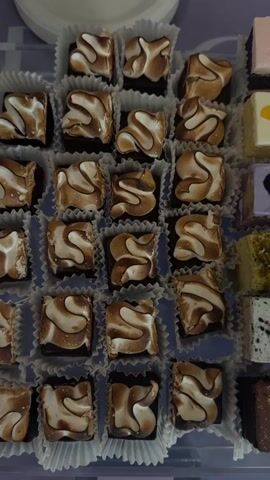 The video displays a collection of cakes, cupcakes, and cookies arranged on a clear tray. The cakes are cut into small squares and decorated with various toppings, including toasted meringue, edible flowers, and nuts. Cupcakes are topped with white frosting and fresh berries. The cookies are hexagon-shaped and decorated with edible flowers and chocolate dots. A large cake sits on a purple cake stand. It is covered in white chocolate shards, resembling fractured glass. The tray and cake stand sit on a table. A beige curtain provides a soft backdrop. The video moves from a close-up of the pastries to a wider shot featuring the larger cake.