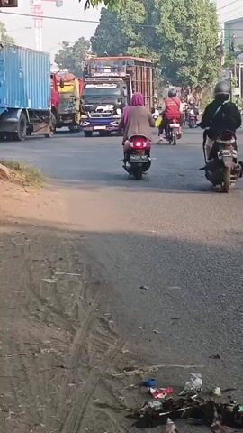 The media file captures a street scene in Kemangsen, Indonesia. The primary subjects are people riding motorcycles. In the foreground, a woman wearing a pink shirt and a hijab is riding on the back of a motorcycle, facing away from the camera. To her right, another person on a motorcycle, wearing a black jacket and helmet, is also visible. The road surface appears to be asphalt, with tire tracks and debris, including trash, scattered on the side.

Further down the road, several other motorcycles are moving in the same direction. A large truck, possibly a cargo truck, is also in motion on the left side of the frame. The setting appears to be a moderately busy street with buildings and trees lining the road. The lighting suggests it is daytime, possibly morning or late afternoon, with soft sunlight casting shadows. The weather appears to be clear.

The overall impression is one of everyday street activity in a developing area, characterized by informal transportation and a visible amount of litter. There is no clear indication of a specific event, but rather a snapshot of daily life. Some text is visible on the side of the truck, but it is not legible. The scene is devoid of any overt emotional expression, presenting a neutral observation of the environment.