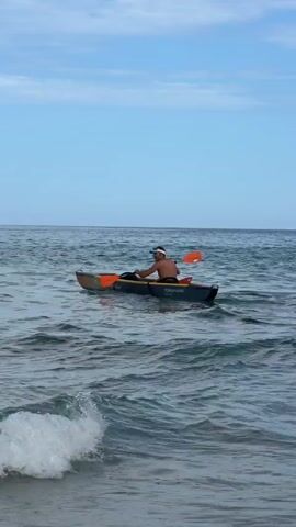 A man wearing a white cap is kayaking in the ocean. The water is choppy with waves breaking in the foreground. The sky is blue with some clouds. The man is holding an orange paddle and appears to be paddling away from the camera.
