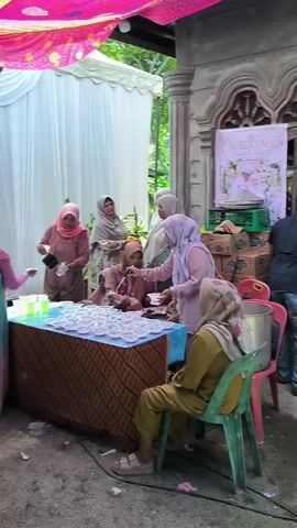 The video shows a group of women, all wearing headscarves, gathered around a table laden with small bowls. One woman in a pink headscarf and a dusty rose colored dress is pouring a green liquid from a bottle into cups. Another woman, also in a pink headscarf and a teal and pink dress, stands next to her. To their right, a woman in a lighter pink headscarf and patterned dress is seated behind the table, and appears to be serving or interacting with the people in front. Further to the right, another woman in a cream headscarf and patterned dress is standing. In the foreground, a woman in a yellow headscarf and beige pants sits on a green chair, facing the table. The setting appears to be outdoors, with a tent or canopy overhead decorated with colorful fabric. There are white curtains behind the table, and a building with a decorative facade is visible on the right. The ground is covered in gravel. The overall scene suggests a gathering or event, possibly a wedding reception, where food and drinks are being served.