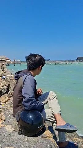 A young man sits on a rocky shore next to a calm, blue ocean. He is wearing a hoodie and light-colored pants, with a helmet resting beside him. In the background, a long pier stretches out over the water, lined with buildings and structures. The sky is clear and blue, indicating a sunny day. The man is looking out at the ocean, appearing relaxed and contemplative.