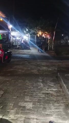 A night scene is depicted, possibly on a street or in an outdoor seating area. The lighting is artificial and dim, with several bright light sources creating lens flares. In the background, there appears to be a food stall or small shop with people standing around it. To the right of the stall, there are trees decorated with hanging lights. A motorcycle is parked or being ridden in the mid-ground. In the foreground, there is a paved area with a textured surface, possibly cobblestone or brick. A white car is seen driving past from left to right. The overall atmosphere suggests a casual evening setting, possibly in a marketplace or a roadside eatery.