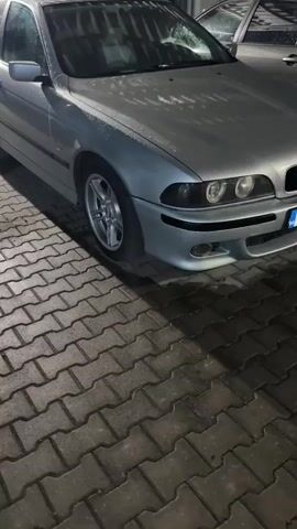 The video displays the front of a silver BMW car, parked outdoors on a paved surface made of cobblestones. The car's hood, windshield, and parts of the body are covered in water droplets, suggesting recent rain or current light precipitation. The scene appears to be at dusk or night, indicated by the artificial lighting reflecting on the car and the general darkness of the background. Another vehicle is partially visible behind the BMW. The camera slowly pans from the driver's side front wheel towards the center of the car's front, focusing on the headlights, grille, and bumper. The Bulgarian license plate "EB 4556 BH" is clearly visible on the front bumper. The blue "BG" country code is also visible on the left side of the license plate. No people are present in the video.