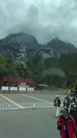 A group of people are standing and walking around a paved area with a white fence and yellow markings on the ground. In the background, there are mountains covered in clouds, trees, and some buildings, including a green tourist bus and a tower with antennas. Several signs with red lettering are visible. The weather appears overcast.