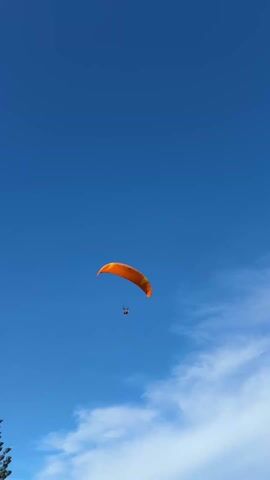The video shows a paraglider in the sky. The paraglider is orange with yellow accents and a single occupant. The sky is mostly blue, with a few white clouds visible in the lower right corner of the frame. There is a hint of green foliage in the bottom left corner, indicating the presence of trees. The camera angle is looking upward.