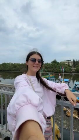 A young woman with dark hair in a braid, wearing sunglasses, a pink sweatshirt, and a white crossbody bag, takes a selfie video on a bridge. She is smiling and looking at the camera. Behind her, several colorful fishing boats are docked in a shallow body of water lined with trees. The sky is cloudy.