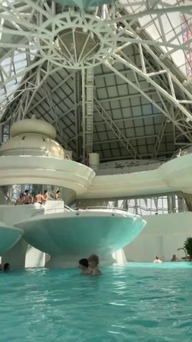 The video shows the interior of a large, modern water park. People are swimming and standing in a large pool of turquoise water. Above them is a complex structure featuring rounded, white platforms that appear to be partially submerged in the water. People are also on these platforms, some standing and some sitting. Above this is a large, white, dome-like structure, and above that, a complex metal framework forming a grid-like roof with glass panels. The environment appears bright and airy due to the large windows.