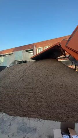 A dump truck is unloading a large pile of sand. The truck's bed is raised high, and sand is pouring out of it onto the ground. Two men are standing near the pile of sand, seemingly overseeing the unloading. In the background, a brick building with windows and a white railing is visible. The sky is clear and blue. The scene appears to be an outdoor construction or landscaping site.