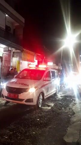 Here is a description of the video:

The video shows an ambulance parked on a city street at night. The ambulance is white with red stripes and the word "AMBULANCE" written on the side. It has its red lights flashing on the roof. There are several people standing around near the ambulance, and some are looking at the camera. The street is wet and dimly lit by streetlights. There are buildings visible in the background. The ambient sound includes someone reciting a religious text.
