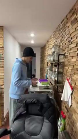 A young man wearing a black beanie and a light blue hooded sweatshirt stands at a small table in a well-lit room. He's picking up what appears to be dried herbs from a small plate and placing them onto a digital scale. Behind him, a wall is covered with a brick-like design. A three-tiered shelf is mounted on the wall and is lined with glass jars filled with various herbs. A black jacket is draped over a chair in the lower left of the frame.  Several bottles of cleaning supplies are visible on the floor in the lower right. The setting seems to be indoors, possibly a kitchen or utility room.