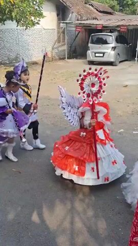 The video shows a group of children participating in a parade. They are dressed in various costumes. Some children are wearing drummer uniforms with red jackets, white pants, and red hats, and they are carrying drums and drumsticks, marching in rhythm. Other children are dressed in more elaborate costumes, including one in a purple dress with a feathered headdress and another in a red and white outfit with large wings and a tall headdress. A woman in a red hijab and a floral shirt is walking with the children, seemingly leading or supervising the parade. The parade is taking place on a paved road or path, with buildings and trees visible in the background. There are also some vehicles parked in the background. The overall scene depicts a festive event likely for children.