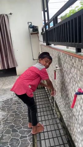 A young boy, wearing a red shirt, black pants, and a black skullcap, stands outside a building and washes his hands under a faucet. The scene appears to be in a warm climate, indicated by the boy's attire and the open-air setting. He is barefoot on a tiled floor with a metal grate in front of him, which allows water to drain away. Behind him, a wall is covered in a mosaic of small, light-colored tiles. A dark-colored railing runs along the top of the wall. To the left, a beige curtain hangs beside a white wall, and a blue water pump is mounted on the wall. Two faucets are visible; one is silver with a red handle, and another is partially obscured and has a blue handle. The boy is smiling and seems to be enjoying himself as he washes his hands, with water splashing around his hands and arms. The lighting suggests it is daytime, possibly morning or afternoon. There is no visible text in the video.