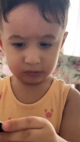 A close-up shot of a toddler is featured. The toddler, wearing an orange tank top with a crab pattern, is looking down and concentrating on an object in their hands. The toddler's head is tilted slightly, and their eyebrows are furrowed, suggesting focus or mild concern. In the foreground, the toddler's hands are holding a small, dark-colored object, possibly a puzzle piece or a toy. Below the toddler's hands, a blue silicone mold with various shapes, resembling cookie cutters or ice cube molds, is visible. The background is out of focus, showing a patterned surface, likely a table or tablecloth, and a hint of what appears to be a curtain or blinds. The lighting suggests an indoor setting with natural light coming from the side. The audio includes a child's voice saying "Misha" and then asking "Show me."