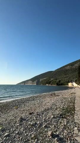 The video presents a serene coastal scene on a clear, sunny day. It opens with a wide shot of a pebble beach, where small, light-colored stones cover the ground. The calm, blue-green sea stretches out to the left, exhibiting gentle waves. In the distance, a large, green-covered mountain or cliff rises steeply from the water's edge, dominating the right side of the frame. A small wooden bench is visible on the beach, facing the sea.

As the video progresses, the camera slowly pans left, revealing more of the extensive coastline and the imposing mountains that hug the shore. The water appears tranquil, reflecting the bright blue sky above, which is completely devoid of clouds. The mountains are densely forested with lush green vegetation, with some exposed rock formations visible near the waterline. A faint, distant object, possibly a small boat, can be seen on the water. The overall atmosphere is peaceful and natural, showcasing the rugged beauty of the coastal landscape in Bratica - Braticë, Montenegro. There is no visible text in the video.