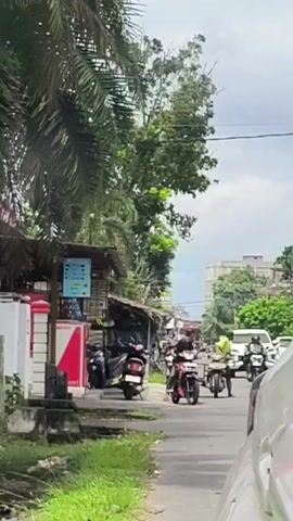 A video shows a street scene with several motorcycles and a white car. On the left side of the road, there is a small stand with a sign that has blue and white text. A red and white refrigerator is visible behind the stand. A palm tree with large fronds is in the foreground on the left. Several motorcycles are parked in front of the stand. Two motorcycles are on the road, one red and one white, with riders. A person in a green shirt is on a white motorcycle to the right of the red motorcycle. A white SUV is visible in the background on the right side of the road. The sky is overcast.