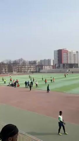 The video shows a group of people playing soccer on a green field. Some are wearing red vests, some are wearing neon yellow tops. Several individuals stand along the edge of the field. The setting appears to be an outdoor sports complex or school, with buildings visible in the background. The sky is slightly overcast.