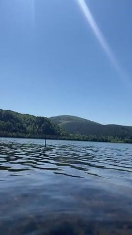 The media file displays a serene natural landscape, likely a lake or reservoir, with forested hills in the background. The camera is positioned very low, almost at water level, capturing the gentle ripples on the water's surface in the foreground. A thin, dark, stick-like object protrudes from the water slightly left of center. Beyond the water, a verdant hillside covered in trees rises, with a lighter green patch possibly indicating a clearing or different type of vegetation. Further in the distance, more hills stretch across the horizon. The sky is clear and bright blue, suggesting a sunny day, likely morning or early afternoon. A strong sunbeam is visible in the upper left corner, casting a bright streak across the sky. The overall scene conveys a sense of calm and natural beauty. No people, animals, or visible text are present. Given the context, this could be the Krasnoyarsk Reservoir near Sliznevo, Russia.