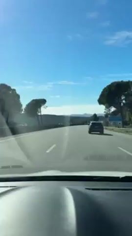 The video depicts a car driving on a highway under a clear blue sky with scattered clouds. Other vehicles are visible on the road ahead. The landscape includes trees lining the roadside and hills in the distance. The perspective is from inside the vehicle, focusing on the road ahead through the windshield. The dashboard of the car is visible in the foreground. The video has a calm and scenic quality.