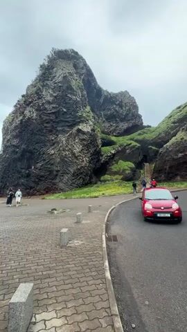 The video shows a cobblestone-paved promenade on a cloudy day. In the background, a large, dark-colored rock formation with green vegetation on top dominates the view. People can be seen walking on a path leading up the side of the rock and along the promenade. A red car is parked on the right side of the screen, next to an asphalt road. The camera slowly pans left to reveal a stony beach and the ocean. Two people in dark clothing are walking towards the beach. A person is sitting on a bench near the shoreline.