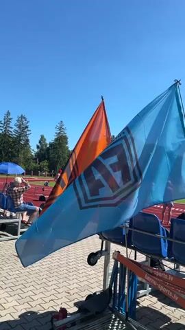 Two flags, one orange and one light blue, are prominently displayed in the foreground, attached to poles that are supported by a metal structure. The light blue flag, positioned in front, displays a logo with the letters "AF" inside a hexagonal shape with a border. Text above the logo on the blue flag reads "FUTBOLA ALIANSE". The orange flag, partially obscured by the blue one, also appears to have a similar logo. In the background, a person wearing a plaid shirt is seated on bleachers. Further back, on a red running track, a person in red shorts and a white shirt is visible. The scene is set outdoors under a clear blue sky, with trees and stadium seating in the distance. A section of orange railing with the text "BERNU-SPORTS.LV" is visible in the lower right corner.