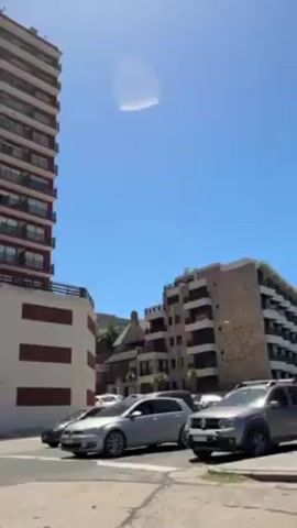 The video depicts a street corner scene on a sunny day. The camera appears to be mounted inside a moving vehicle. 

The scene is dominated by buildings, particularly tall apartment buildings. One prominently displayed building is a high-rise with multiple balconies and a red trim at the top. Adjacent to it is another building, painted in muted blue-grey tones with rounded balconies. There are several other buildings visible in the background, some with more traditional architectural features.

The street is paved and lined with vehicles including a dark-colored hatchback and a silver sedan. The road markings are somewhat faded. There is a traffic sign visible on the side of the road.

The sky is clear blue with no clouds.