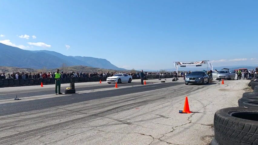 The video shows two cars at a drag racing event. On the left is a white Camaro. On the right is a dark gray BMW sedan. The cars start racing down the track with the Camaro slightly pulling ahead.  Many spectators line the sides of the track, behind black barriers made from tires.  Officials in bright vests stand between the cars. Mountains can be seen in the background under a blue sky with scattered clouds.  Orange cones mark the edge of the race track. A banner reading "DRAG RACING" marks the finish line.