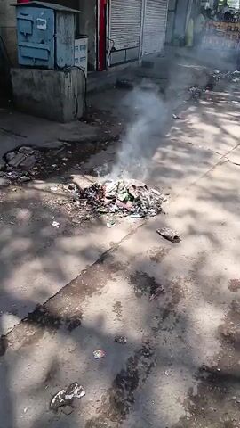 The video shows a burning pile of trash in a street. The pile is made up of various items, including paper, plastic, and other debris. Smoke is rising from the pile, and the surrounding area is littered with additional trash. The street is paved and cracked in places. In the background, there are buildings with closed metal shutters, and people are standing or walking by. A motorcycle is parked nearby. The scene appears to be a street in a populated area, possibly a market or commercial district. The lighting is bright, indicating it is daytime.