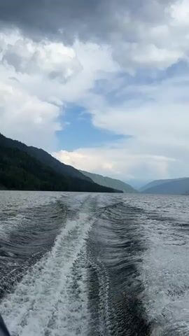 A boat travels across a body of water, leaving a foamy wake behind it. The water is calm with gentle waves. On either side of the water are tree-covered mountains under a partly cloudy sky. The sky is a mixture of dark grey clouds and patches of bright blue sky. The mountains on the left are darker and more densely forested than the mountains on the right, which appear lighter and more distant. The camera is positioned at the back of the boat, looking forward.