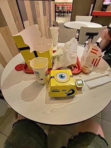 The video shows a table in a fast-food restaurant. On the table are several KFC items, including a box decorated with Minions imagery, a burger, french fries, and drinks. A person's hand enters the frame and opens the Minions-themed burger box, revealing the burger inside, which also features a Minion design on the bun.