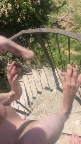 The video shows a close-up of a hairless cat's hind legs and paws as it urinates. The cat is positioned on a concrete spiral staircase, with green foliage and trees visible in the background. The cat's paws appear dirty, and it is holding one paw up, revealing its claws. The camera angle is from above, looking down at the cat.